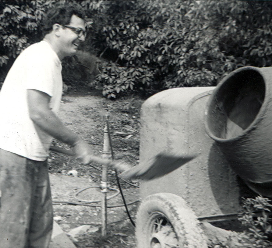 Man with glasses and white t-shirt using flat-nosed shovel to pitch sand and cement into a turning cement mixer. Image of Michael Clarke Rubel at work building his Castle. Ca. 1969.